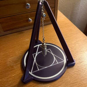 May include: A purple pendulum stand with a silver pendulum hanging from the top. The stand has a circular base with a geometric design.
