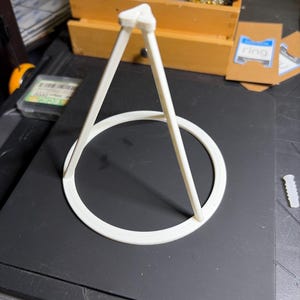 May include: A white, three-dimensional printed stand featuring a circular base with three angled support arms that converge at a triangular top point. The structure is constructed from a solid, matte-finish material and is designed to hold objects.