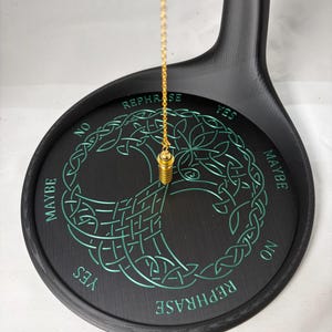 May include: A black divination pendulum board with a gold chain and weight. The board features a teal Celtic tree of life design and the words "Yes", "No", "Maybe", and "Rephrase".