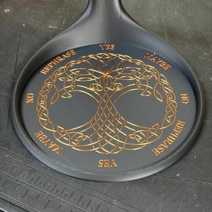 A black circular pendulum stand rests on a dark surface, displaying a gold-colored Tree of Life design and divination words around the rim.
