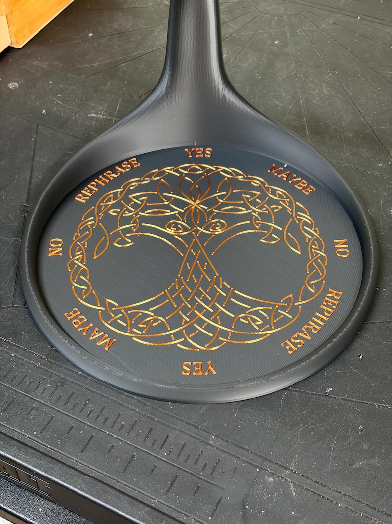 A black circular pendulum stand rests on a dark surface, displaying a gold-colored Tree of Life design and divination words around the rim.