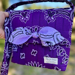 Puede incluir: Un bolso bandolera acolchado con estampado de bandana en color morado oscuro y patrones de cachemira en blanco y negro. El bolso tiene un lazo de tela morado claro en la solapa frontal y una etiqueta rectangular blanca que dice Billye Jo's Designs handmade.