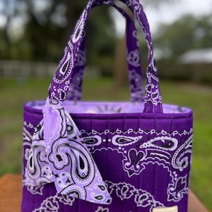 May include: A quilted tote bag featuring a vibrant purple and white paisley bandana print. The bag has two matching fabric handles and a decorative knotted fabric tie attached to one side. A small rectangular label is stitched to the bottom corner.