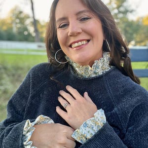 May include: A dark charcoal gray long-sleeved sweater featuring a high ruffled collar and matching ruffled cuffs in a light cream fabric with a delicate blue and yellow floral pattern. The sweater has a textured knit finish.
