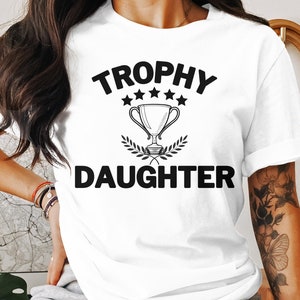 Może przedstawiać: Biała koszulka z czarnym napisem "Trophy Daughter" z grafiką trofeum i pięcioma gwiazdami.