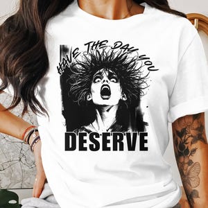 Puede incluir: Camiseta blanca con un gráfico en blanco y negro de una mujer con el pelo de punta. El texto "HAVE THE DAY YOU DESERVE" está impreso en la camiseta.