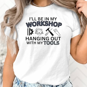 May include: White t-shirt with the text "I'll be in my workshop hanging out with my tools". The text is in black with a gray outline. There are black and gray illustrations of tools below the text.