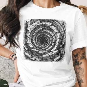 May include: White t-shirt featuring a black and white graphic of a spiral well design. The artwork has a detailed, textured appearance, creating a sense of depth. The shirt is made of a soft material.