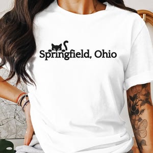 Puede incluir: Camiseta blanca con un gato negro asomándose detrás del texto "Springfield, Ohio".
