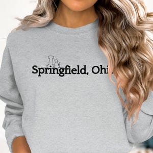 Puede incluir: Una sudadera gris con un gráfico negro de un perro, un gato y un pájaro con el texto "Springfield, Ohio" impreso en la parte delantera.