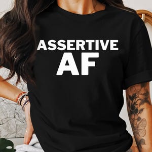 May include: Black t-shirt with the white text "ASSERTIVE AF". The shirt has short sleeves and a crew neck. The text is in a bold, sans-serif font.