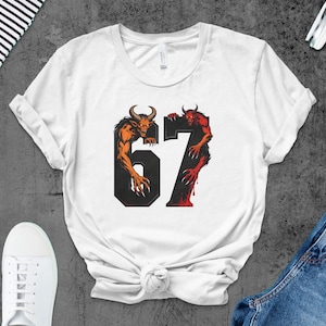 May include: White t-shirt featuring the number 67 in a black font, with orange and red demon illustrations. The shirt is styled with white sneakers and denim jeans. The design is centered on the front of the shirt.