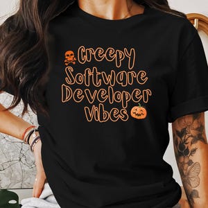 Op de afbeelding: Zwart T-shirt met oranje tekst die "Creepy Software Developer Vibes" luidt met een schedel en gekruiste beenderen en een pompoenafbeelding.