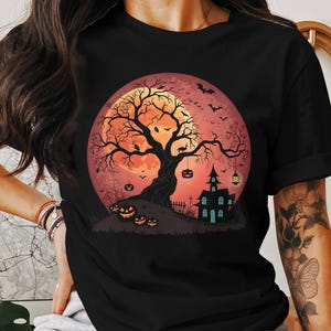 May include: Black t-shirt featuring a Halloween design. The graphic includes a large orange moon, a spooky tree, bats, a haunted house, and jack-o'-lanterns. The design is set against a gradient background.