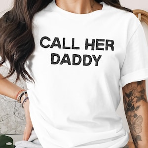 Puede incluir: Camiseta blanca con el texto "CALL HER DADDY" en letras negras en negrita. La camiseta tiene mangas cortas y cuello redondo. La imagen muestra la camiseta en una persona.