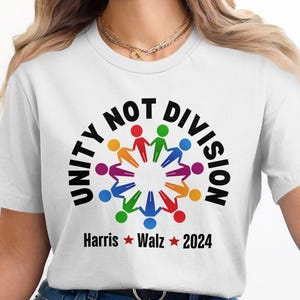 Może przedstawiać: Biała koszulka z czarnym kołem graficznym, na którym widnieje napis "Unity Not Division" oraz koło kolorowych ludzików trzymających się za ręce. Pod grafiką znajduje się tekst "Harris * Walz * 2024".