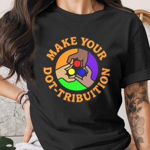Może przedstawiać: Czarny t-shirt z kolorową grafiką. Projekt przedstawia dłonie trzymające czerwone, żółte i niebieskie kropki w okrągłej, tęczowej obwódce. Tekst brzmi "MAKE YOUR DOT-TRIBUTION".