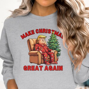 May include: A gray sweatshirt with a red graphic print of a man sitting in a chair with a Christmas tree behind him. The text on the sweatshirt reads "Make Christmas Great Again".