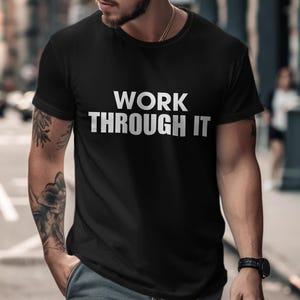 May include: Black t-shirt with the white text "WORK THROUGH IT". The shirt is short-sleeved and appears to be made of a soft material. The image is taken outdoors, with a blurred background of a city street.