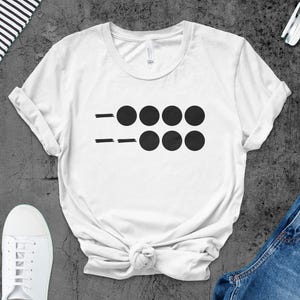 Puede incluir: Camiseta blanca con un diseño gráfico negro. El diseño presenta dos líneas horizontales con cuatro círculos negros arriba y abajo. La camiseta se combina con zapatillas blancas y vaqueros azules.