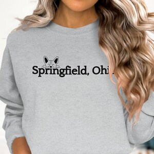 Puede incluir: Una sudadera gris con un gráfico negro de un perro asomándose sobre el texto "Springfield, Ohio".