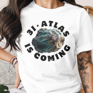 May include: White t-shirt with the text "3I ATLAS IS COMING" in black, surrounding a colorful, textured rock graphic. The shirt is a casual style, suitable for everyday wear.