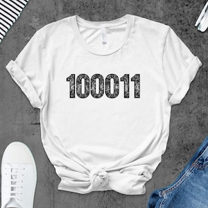 Könnte beinhalten: Weißes T-Shirt mit der schwarzen Aufschrift "100011". Das Shirt ist mit hochgekrempelten Ärmeln und einem Knoten am Saum versehen. Das Bild zeigt weiße Turnschuhe, Jeans und Kopfhörer.