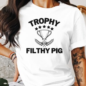 May include: White t-shirt with black text that reads "Trophy Filthy Pig" and a graphic of a trophy with five stars above it.