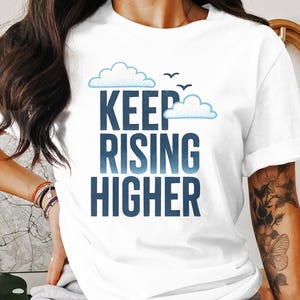 May include: White t-shirt featuring the phrase "KEEP RISING HIGHER" in a gradient blue font, with cloud illustrations and three small birds. A casual wear item.