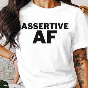 May include: White t-shirt with the words "ASSERTIVE AF" printed in bold black letters. The shirt has short sleeves and is being worn by a person. The person has dark hair and tattoos on their arm.