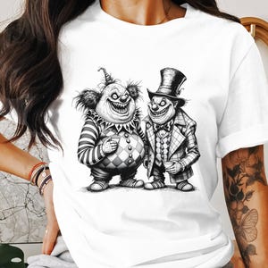 May include: White t-shirt featuring a black and white illustration of two grinning clowns. One clown wears a striped shirt and diamond-patterned pants, the other a checkered suit and top hat. The design is a detailed, cartoon-style graphic.