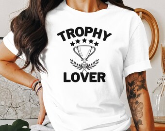 Könnte beinhalten: Weißes T-Shirt mit schwarzem Text "Trophy Lover" und einer Grafik eines Pokals mit fünf Sternen darüber.