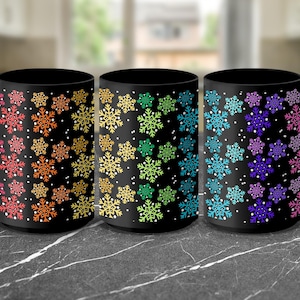 May include: Three black ceramic mugs with snowflake designs in rainbow colors. Each mug features a different color scheme: red to orange, yellow to green, and blue to purple. White dots are scattered around the snowflakes.