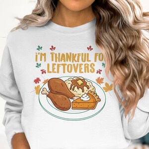 May include: White sweatshirt with the text "I'M THANKFUL FOR LEFTOVERS" in gold and green. A plate with a cartoon illustration of a Thanksgiving meal, including turkey legs, mashed potatoes, and pumpkin pie.
