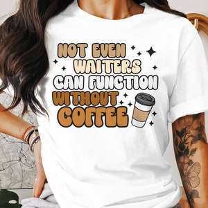 Puede incluir: Camiseta blanca con el texto "NOT EVEN WAITERS CAN FUNCTION WITHOUT COFFEE" en marrón, beige y blanco. Un gráfico de taza de café está al lado del texto. La camiseta tiene mangas cortas.