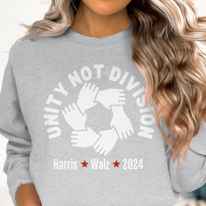 Può includere: Felpa grigia con una grafica bianca che dice "Unity Not Division" con un cerchio di mani che si tengono. Il testo "Harris Walz 2024" è sotto la grafica.