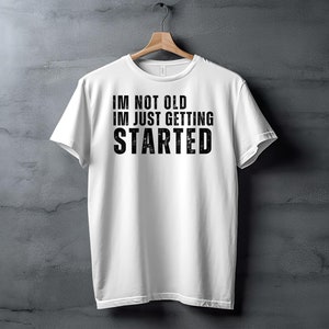 May include: White t-shirt with black text that reads "I'M NOT OLD I'M JUST GETTING STARTED"