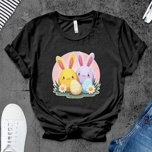 Peut inclure: T-shirt noir avec un motif de deux lapins de dessin animé tenant des œufs de Pâques. Les lapins sont jaunes et violets, et les œufs sont jaunes et bleus.