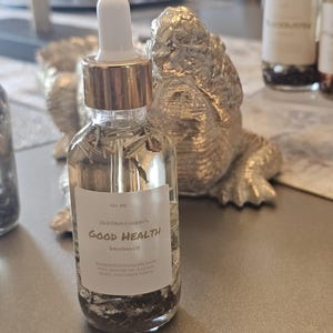 May include: A clear glass bottle with a gold lid and a white dropper top. The bottle is filled with a clear liquid and contains herbs and other natural ingredients. The label reads "Rich Bitch Conjure's Good Health Intention Oil".