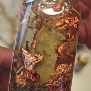 Könnte beinhalten: Eine klare Glasflasche gefüllt mit einer goldenen Flüssigkeit, die Kupferflocken, getrocknete Kräuter und ein großes grünes Blatt enthält. Die Flasche hat einen goldenen Deckel.