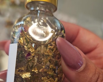 May include: A clear glass bottle with a gold lid filled with a golden liquid and gold flakes. The bottle is held in a hand with a light pink nail polish.