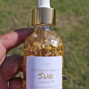 Puede incluir: Una botella de vidrio transparente de Sun Conjure Oil de Rich Bitch Conjure, con una etiqueta blanca y detalles dorados. La botella contiene copos de oro y un cuentagotas. La etiqueta dice "Blessed Intention Oil made with almond oil, natural herbs, and other curios."