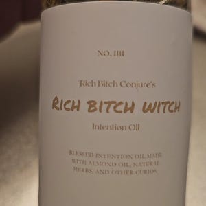May include: A glass bottle with a white label featuring the text "RICH BITCH WITCH Intention Oil." The label also includes the text "Blessed Intention Oil made with almond oil, natural herbs, and other curios."