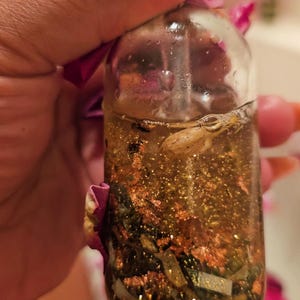 May include: A small glass bottle filled with a golden liquid and gold flakes. The bottle is held in a hand and there are pink flower petals in the background.