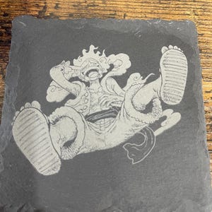 May include: A square, black slate coaster featuring a detailed etched illustration of a cartoon character in a dynamic pose. The character is depicted with outstretched limbs and a joyful expression. The coaster is designed for use with beverages.