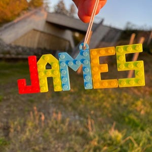Puede incluir: Una colorida etiqueta con el nombre "JAMES" en letras de bloque. Las letras son rojas, amarillas y azules, con una cuerda blanca para colgar. El fondo muestra una escena borrosa al aire libre con hierba y un puente.
