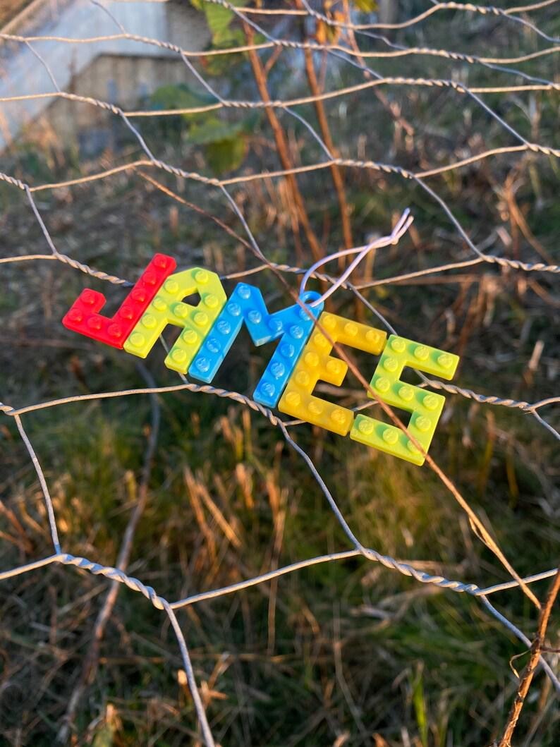 Puede incluir: Una colorida etiqueta con el nombre "JAMES" hecha a mano con bloques de pl&aacute;stico entrelazados. Las letras son rojas, amarillas, azules y verdes, unidas con hilo blanco, sobre un fondo borroso de una valla de alambre y follaje.