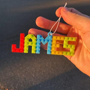Puede incluir: Una colorida etiqueta con el nombre "JAMES" en letras de bloque. Las letras son rojas, amarillas, azules y verdes, conectadas con una cuerda blanca para colgar. La etiqueta con el nombre se sostiene contra un fondo borroso.