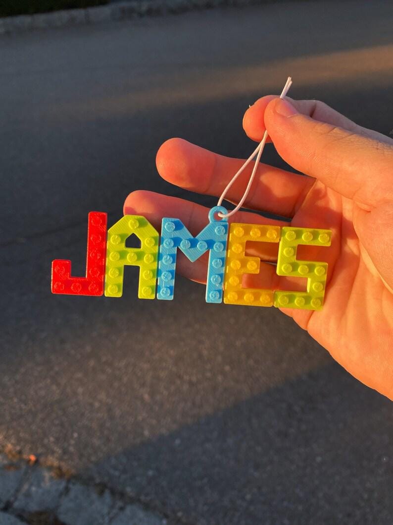 Puede incluir: Una colorida etiqueta con el nombre "JAMES" en letras de bloque. Las letras son rojas, amarillas, azules y verdes, conectadas con una cuerda blanca para colgar. La etiqueta con el nombre se sostiene contra un fondo borroso.