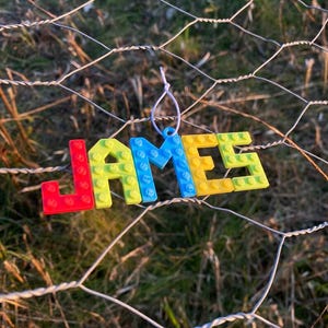 Puede incluir: Un adorno colorido que deletrea el nombre "JAMES" en letras de bloque. Las letras son rojas, amarillas y azules, que se asemejan a bloques de construcci&oacute;n de juguetes. El adorno cuelga de una cuerda blanca sobre un fondo de valla de alambre y hierba verde.